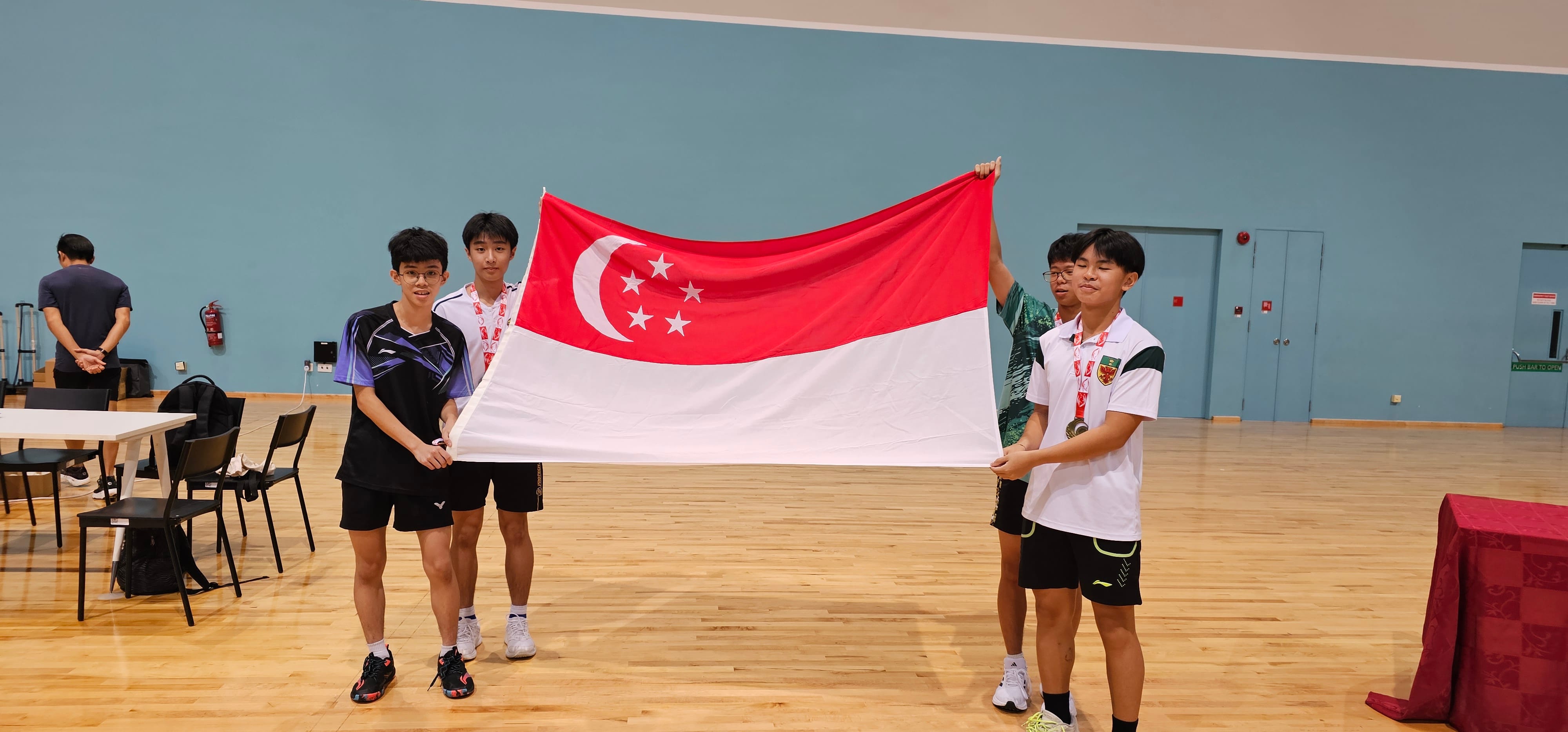 Two students in white uniforms hold up a Singapore national flag on a badminton court.